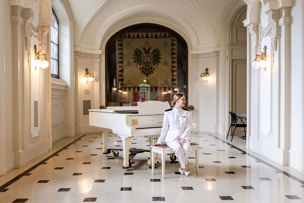 Elegant grand piano in white marble hallway with ornate decor and checkered floor beneath arched ceiling.