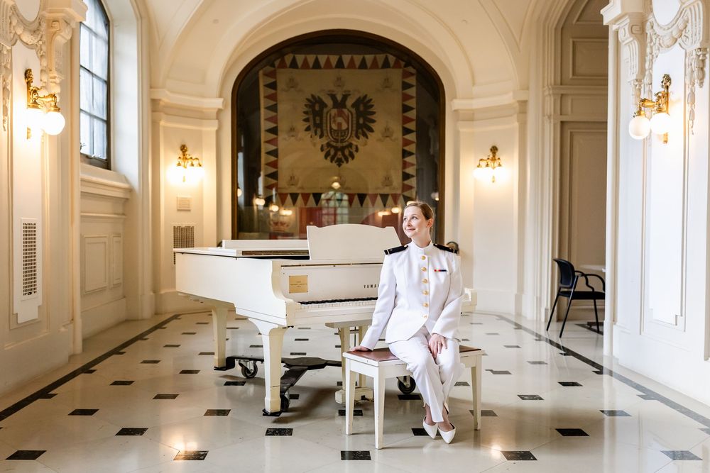 An elegant white grand piano sits in a regal room with ornate walls, checkerboard floor, and golden sconces.