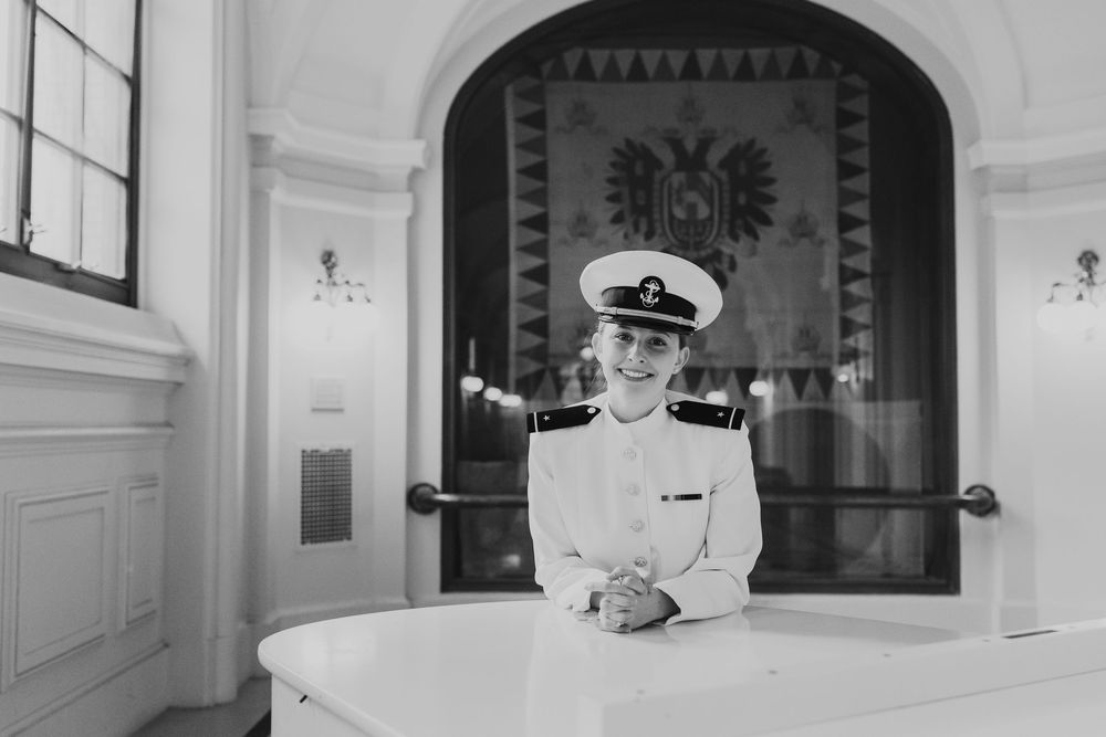 A person in a naval officer uniform sits at a desk in an elegant room with an ornate archway and eagle emblem.