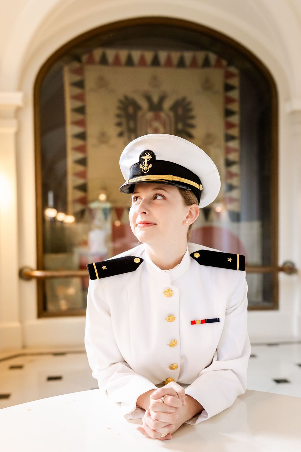 Naval officer in dress whites sits at table under ornate arched ceiling with decorative mosaic.