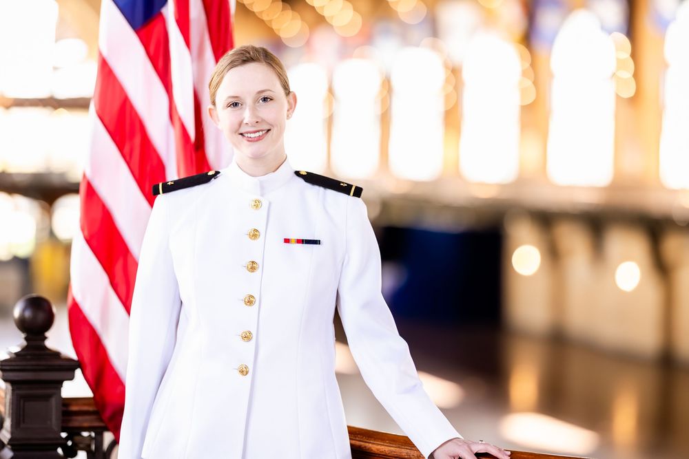 Service member in Navy dress white uniform standing beside American flag in formal setting.