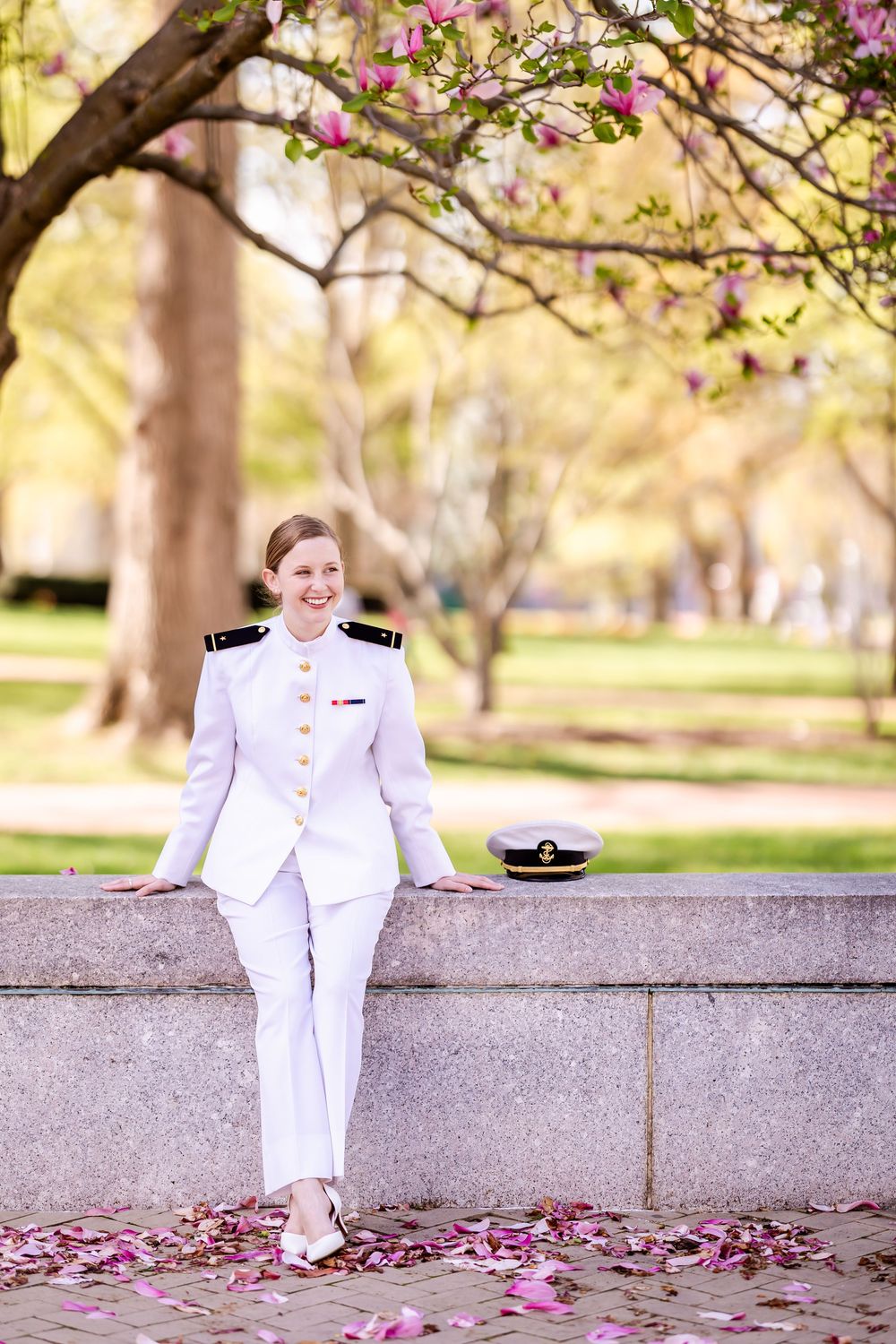 Naval officer in crisp white uniform sitting on stone wall with cherry blossoms falling during spring season.