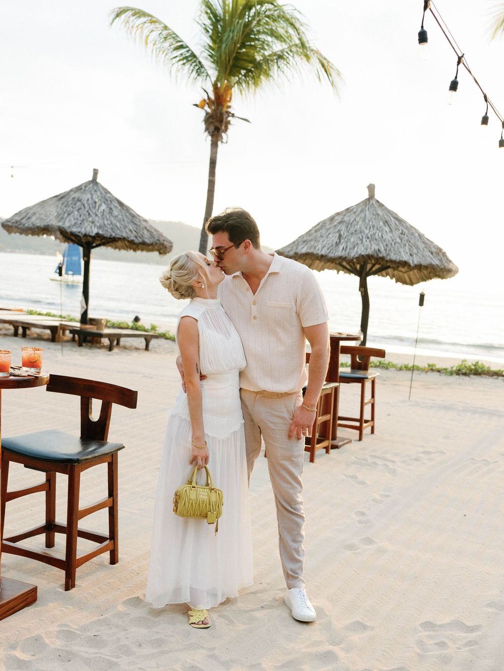 Couple standing on beach near thatched umbrellas and palm trees at tropical resort.