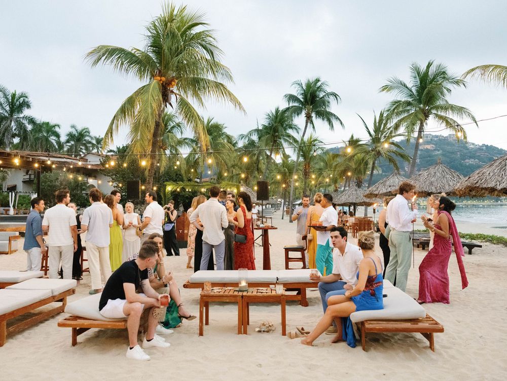 Beach party setup with wooden benches and palm trees as guests mingle during sunset.