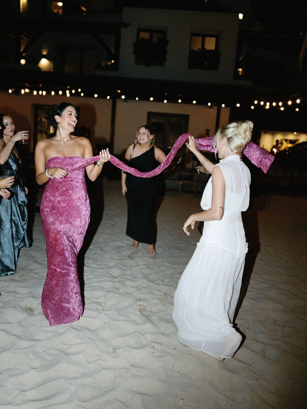 Wedding celebration on sandy beach with guests dancing under string lights at night.