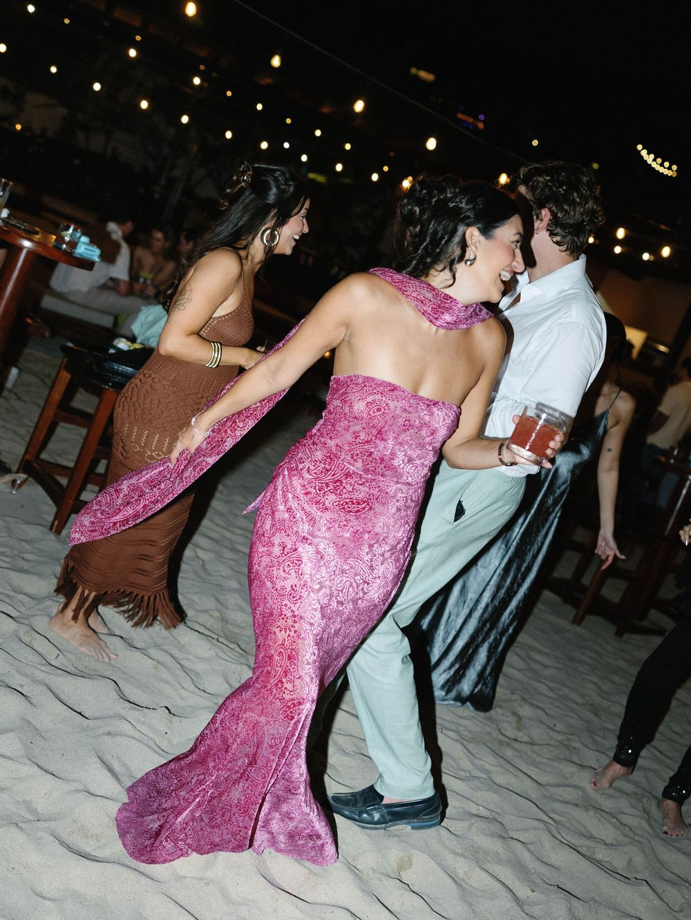 Two people dancing on a beach at night with string lights overhead, one wearing a pink mermaid-style dress.