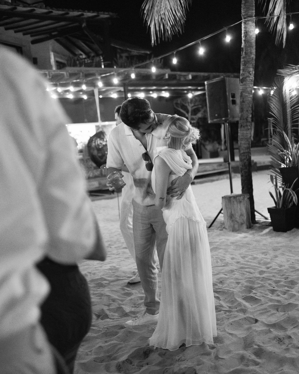 A couple dancing romantically on a beach under string lights near palm trees.