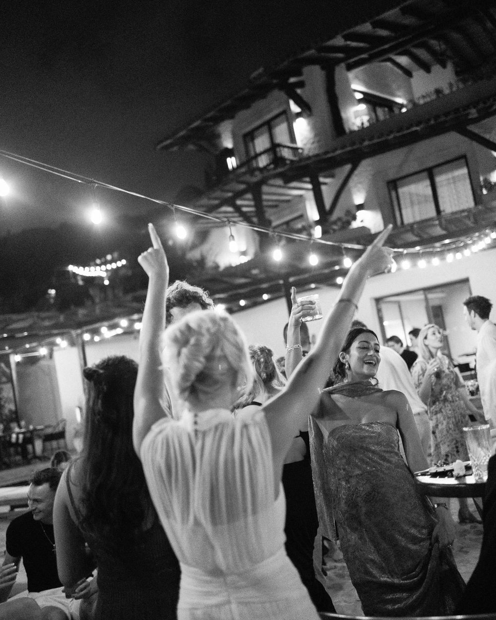 People dancing and celebrating at an outdoor venue with string lights and architecture in the background.