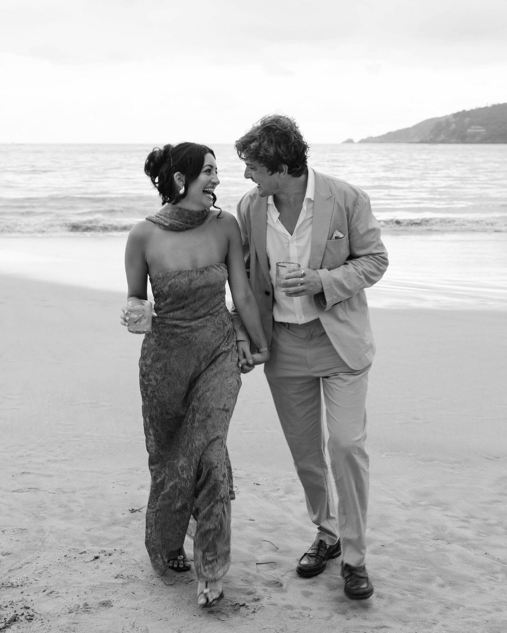 A couple walking and laughing together on a beach with ocean waves in the background.