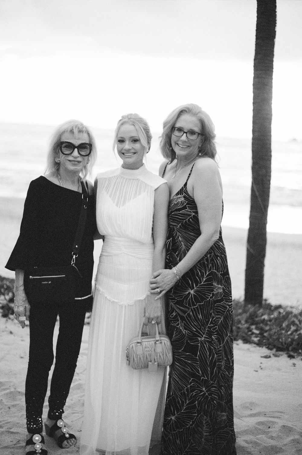 Three women pose together on a beach, with one wearing a white wedding dress.