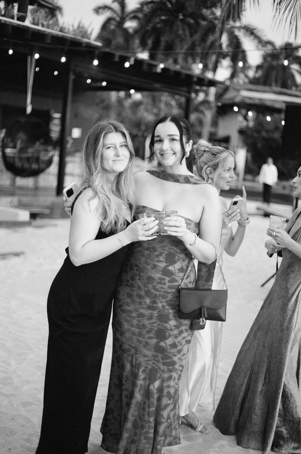 Friends in formal attire sharing drinks and smiles at an outdoor beach event.