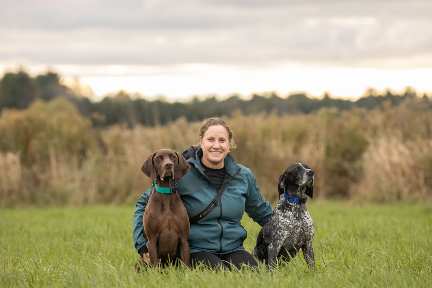 A person in a green jacket sits in a grassy field with a brown and a black spotted dog on either side.