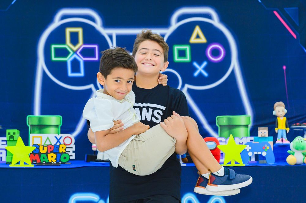 Two children posing together against a blue gaming-themed backdrop with neon controller design and video game decorations.