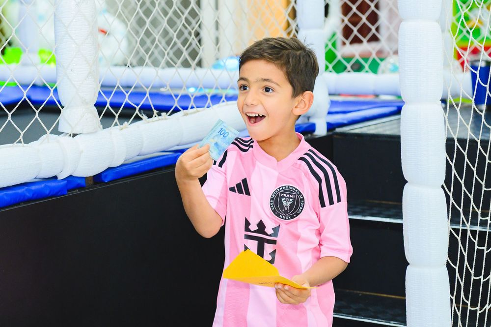Child in pink soccer jersey shows excited expression while standing in front of sports netting.