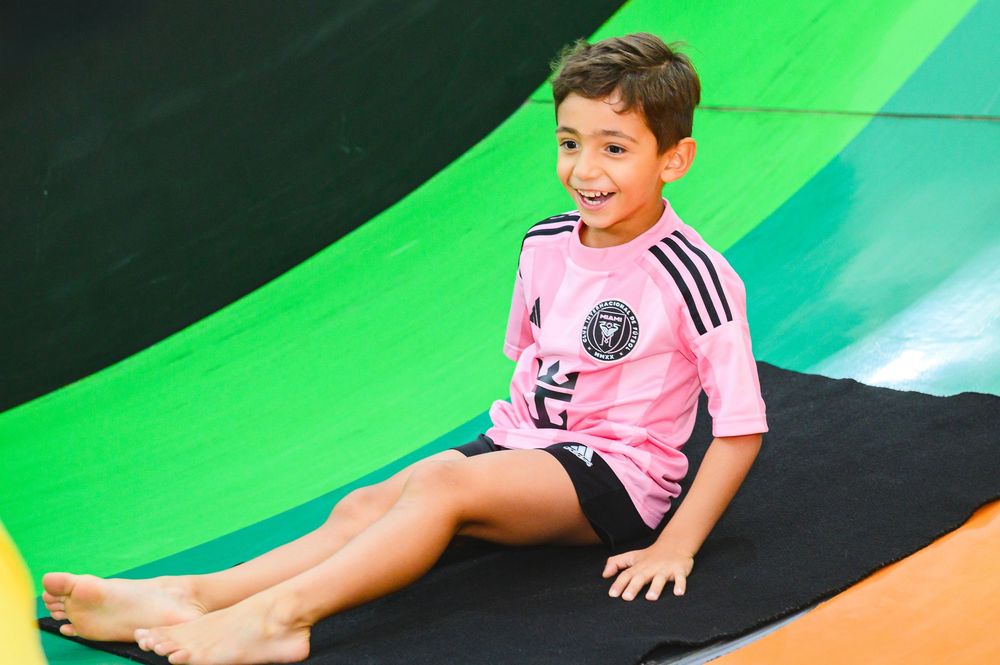 Child in pink sports jersey sitting and smiling on a playground slide with green backdrop.