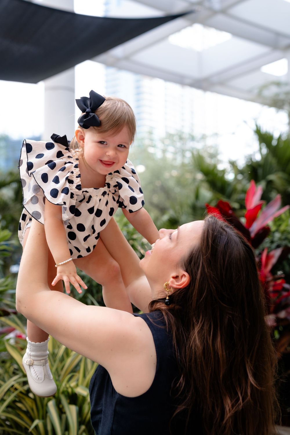 Mother lifts smiling toddler wearing polka dot outfit against tropical garden backdrop.