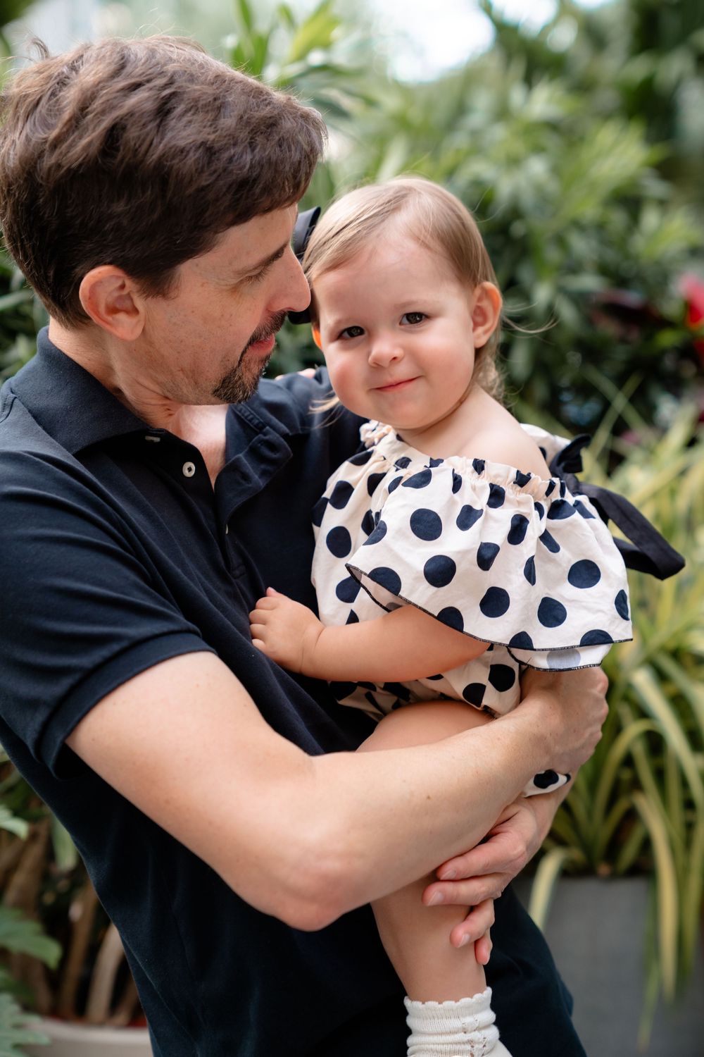 A parent in a navy polo holding a toddler wearing a polka dot dress surrounded by lush greenery outdoors.