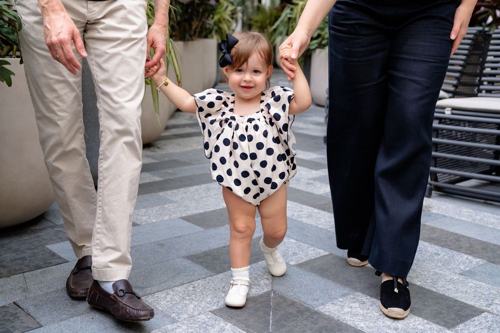 A toddler in a polka dot outfit walks while holding hands with adults on a patterned outdoor path.