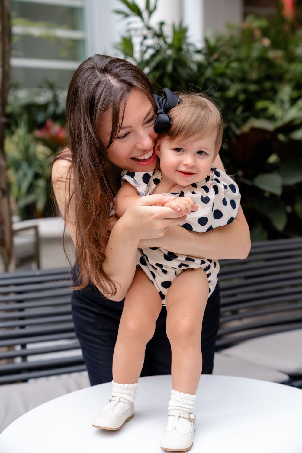 A happy parent holds a smiling toddler wearing a polka dot outfit near green plants outdoors.