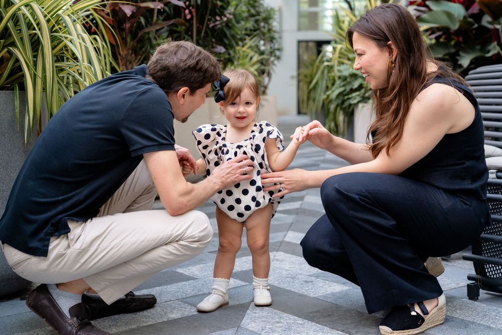 Family interacts with small child wearing polka dot dress in a garden setting with lush plants in background.