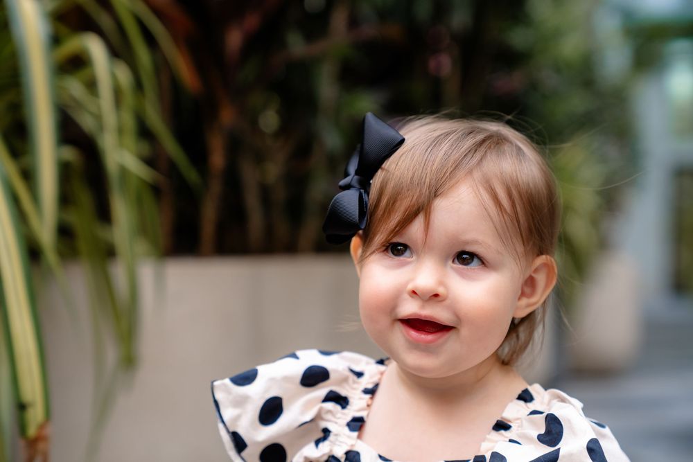 Young child in a polka dot dress with bow smiles at camera against greenery background.