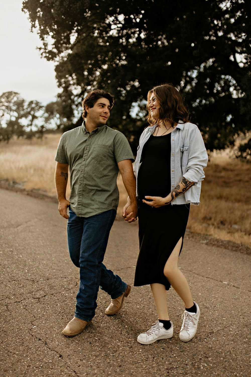 A couple walks hand in hand in Cameron Park with the woman wearing a denim jacket and black dress during a maternity photo shoot.