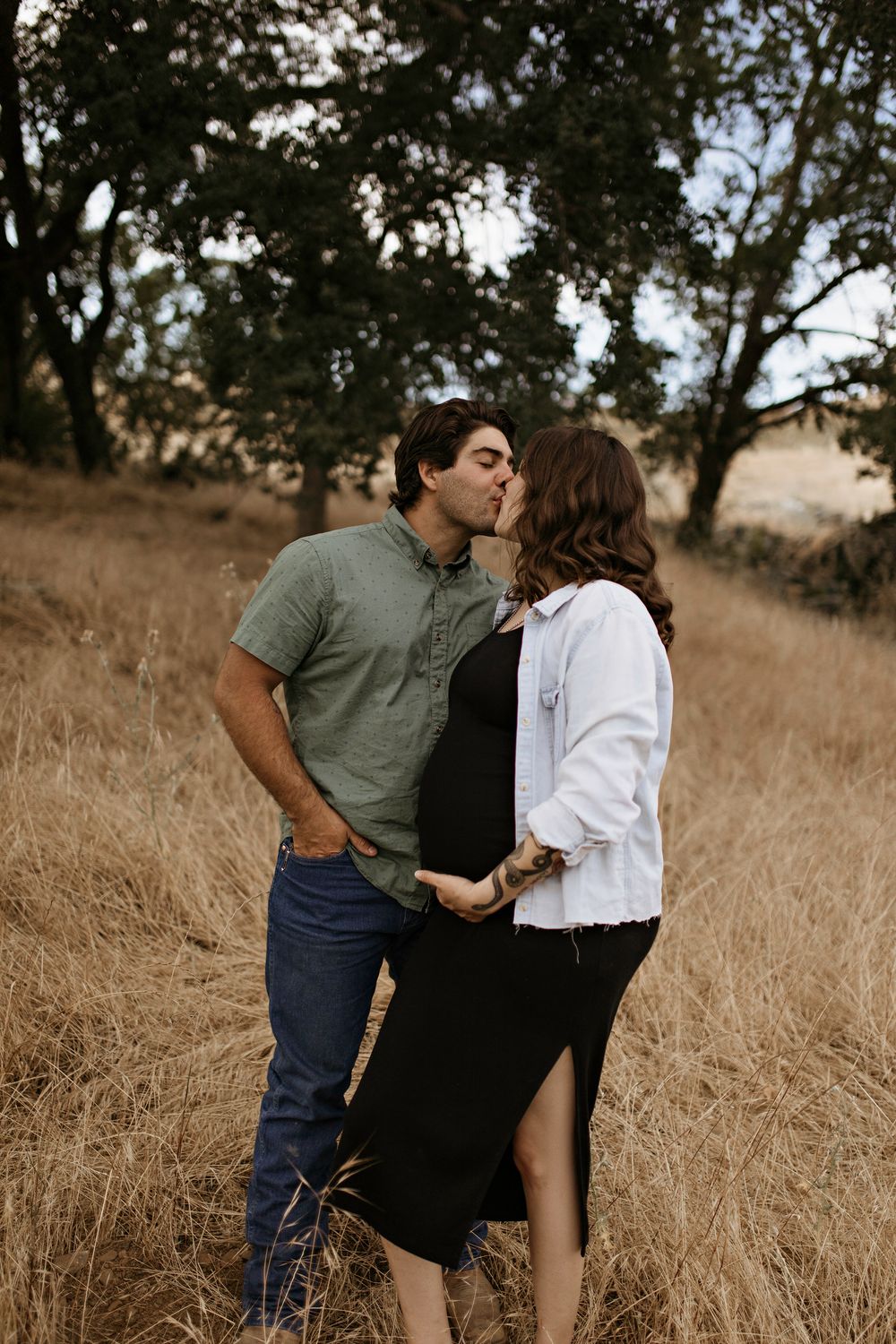 Maternity shoot kiss while standing in tall golden grass with trees in the background during sunset.