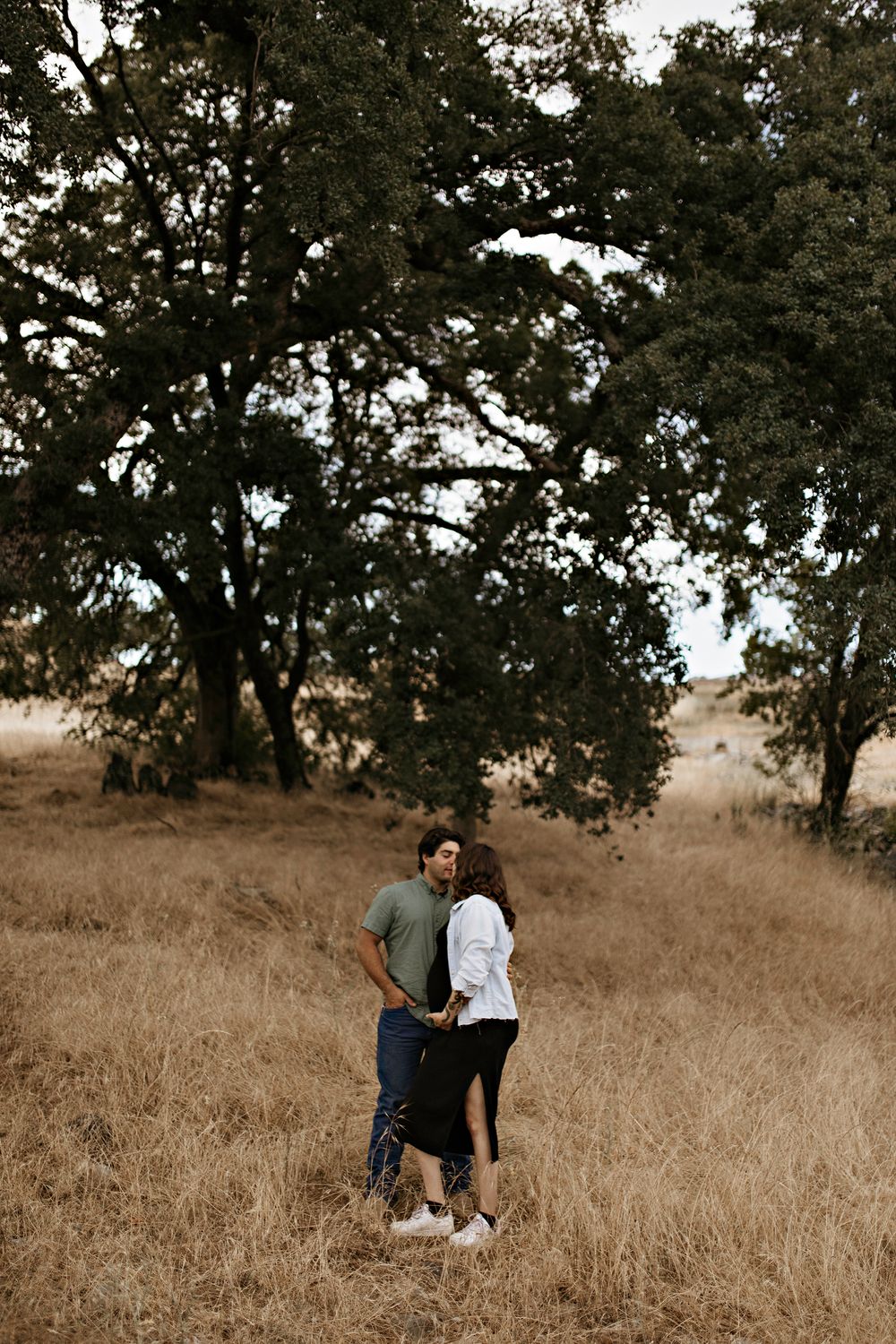 Maternity pose for couple in an intimate embrace among tall golden grass beneath large oak trees in a rustic outdoor setting.