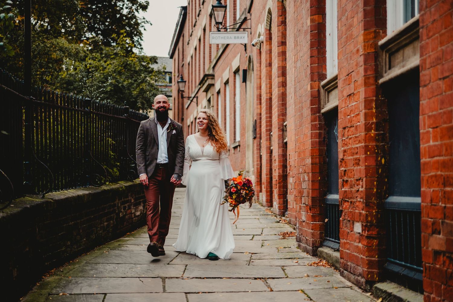 A couple in wedding attire stands together on a brick-lined pathway next to a historic red building.