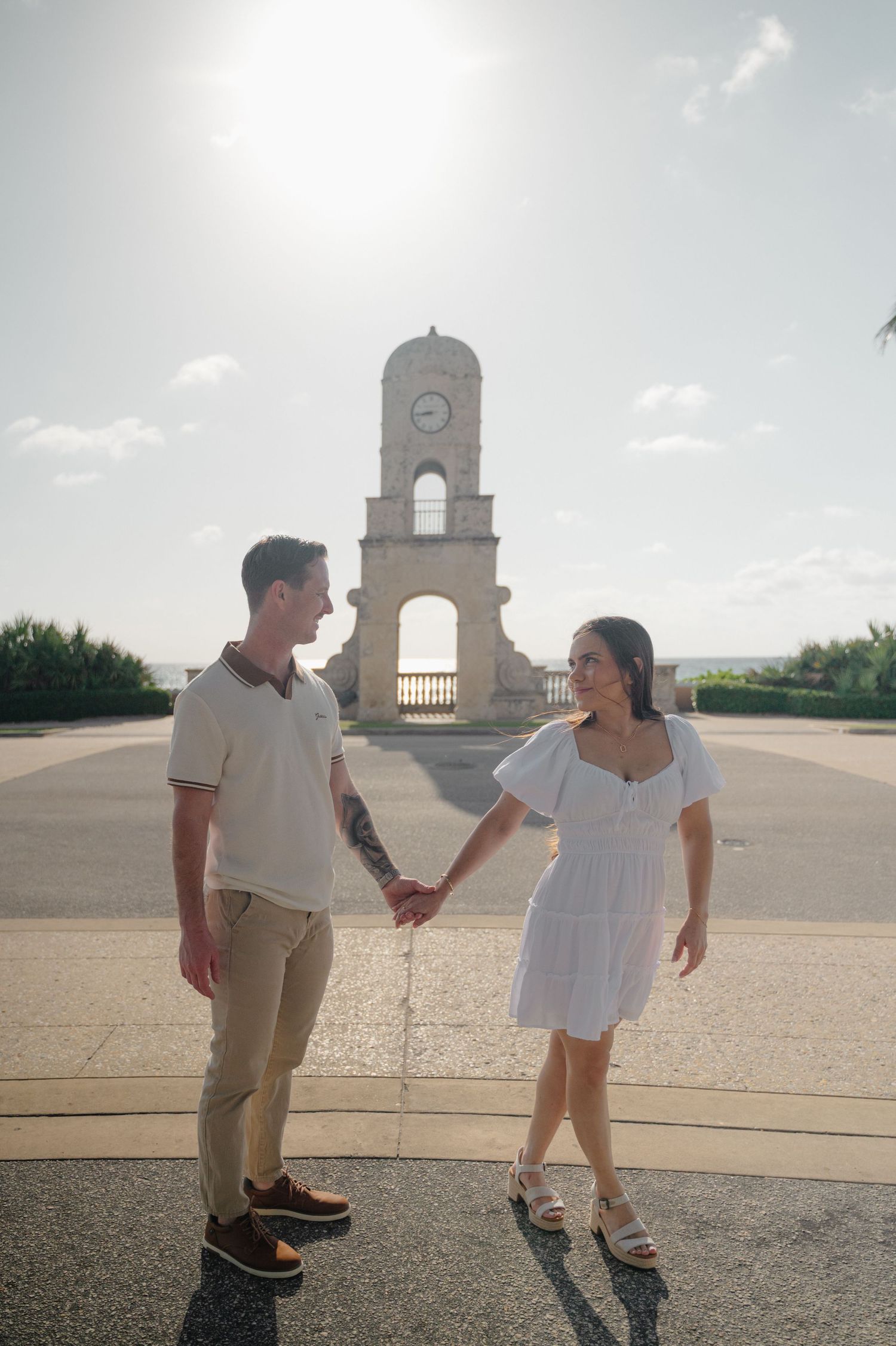 A couple holding hands stands in front of a historic clock tower monument on a sunny day.