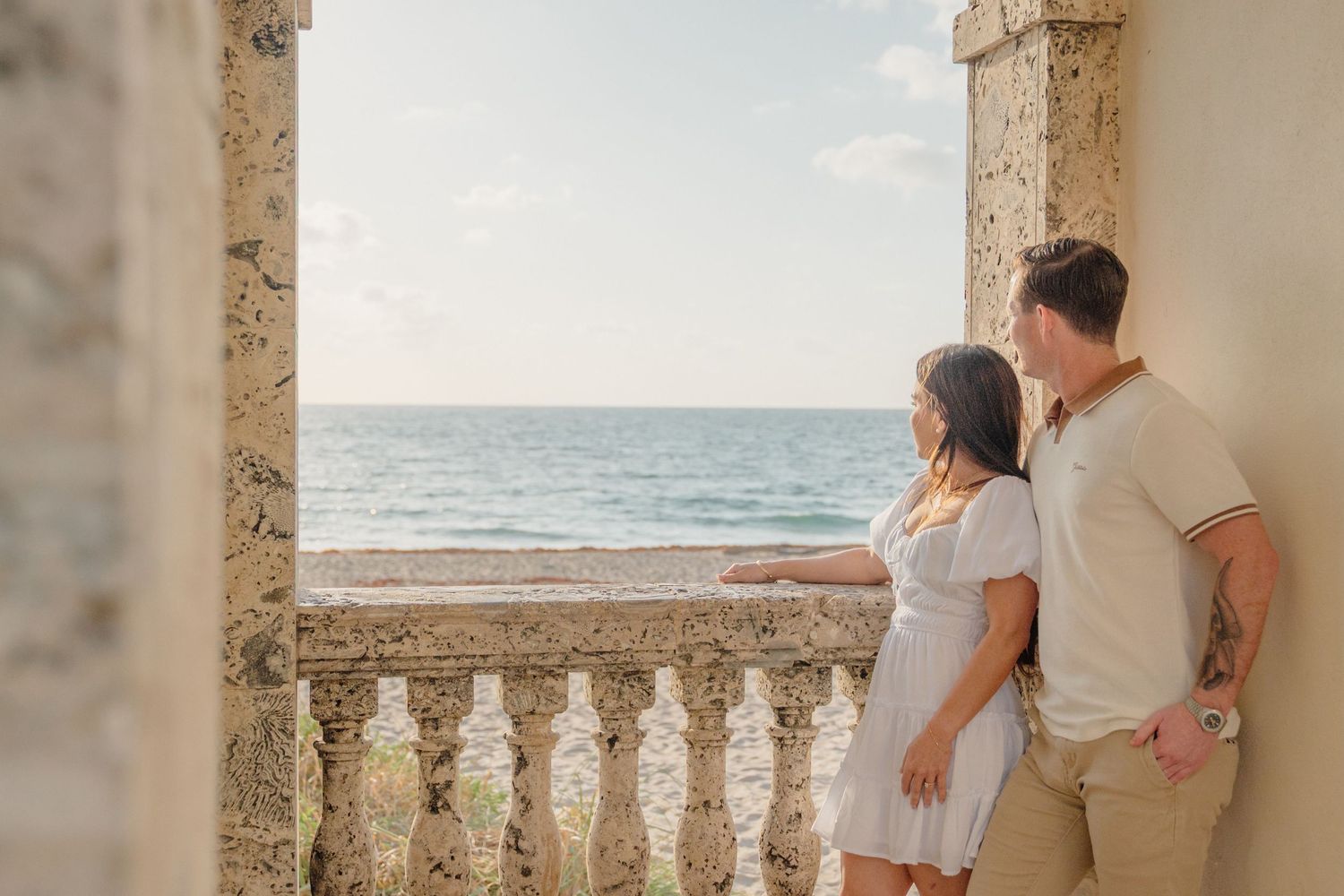 A couple stands at a seaside balcony with ornate stone pillars overlooking the ocean during golden hour.