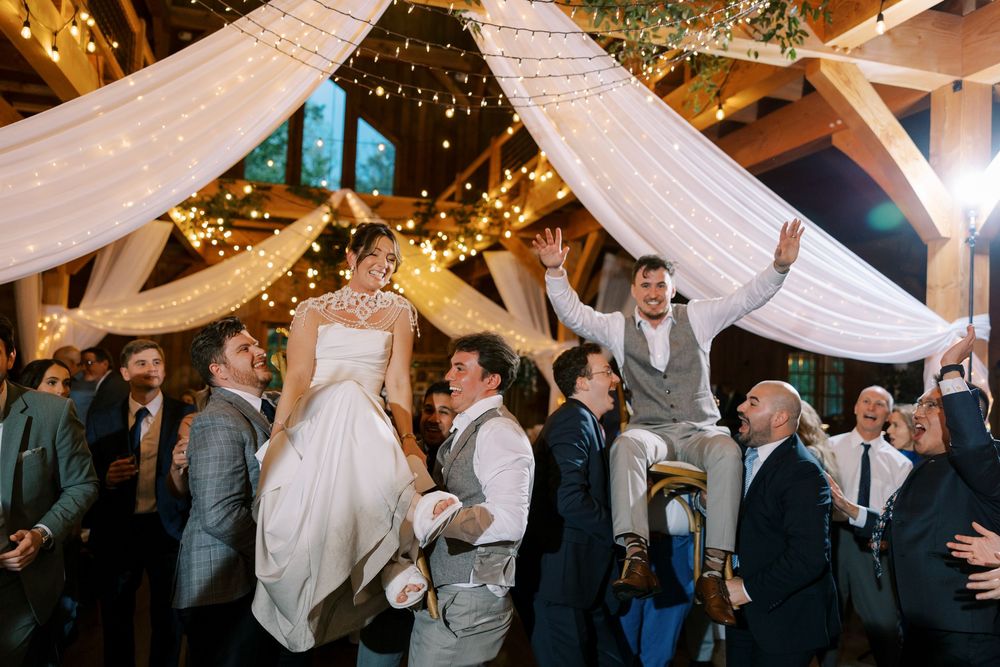 Wedding reception guests lift up celebrants during lively dance celebration under draped fabric and string lights at Windham Manor.