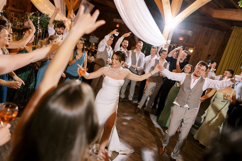 Wedding guests dance and celebrate with arms raised on a rustic dance floor decorated with white drapes at Windham Manor.