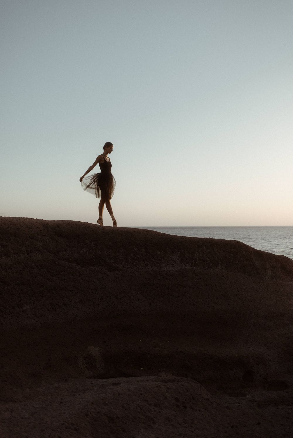 Silhouette of a graceful ballerina posing for a photographer on a cliff edge against an ocean horizon at dusk in La Caleta de Adeje.