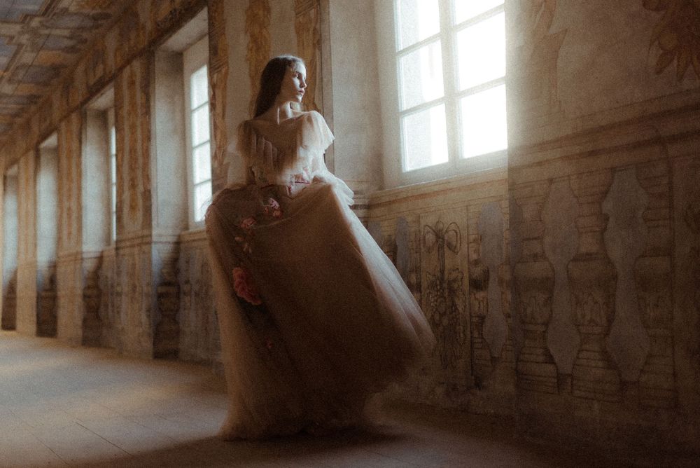 An ethereal figure in a gown sits by ornate windows in a historic hallway bathed in soft natural light. Wedding photographer Tenerife.