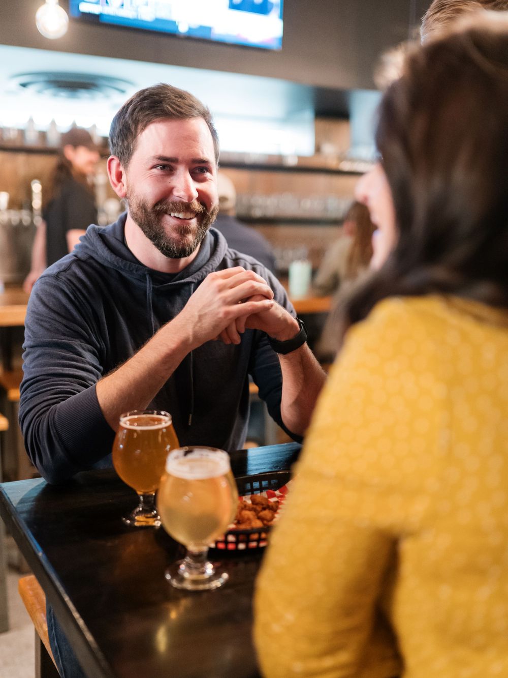 Person in black hoodie engaging in conversation at a bar counter with drinks.