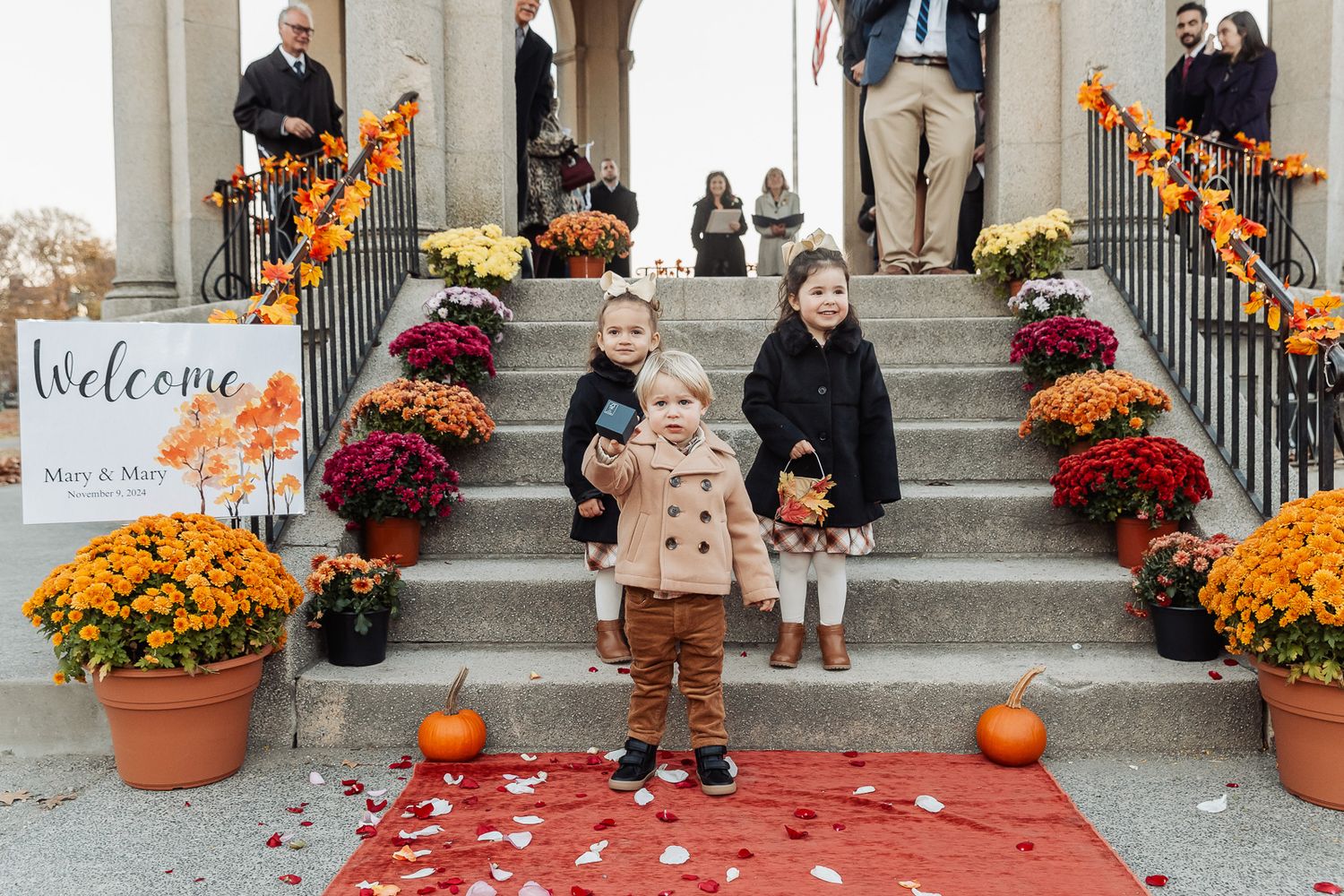 Children and guests gather on decorated steps with orange mums and pumpkins at a fall wedding.