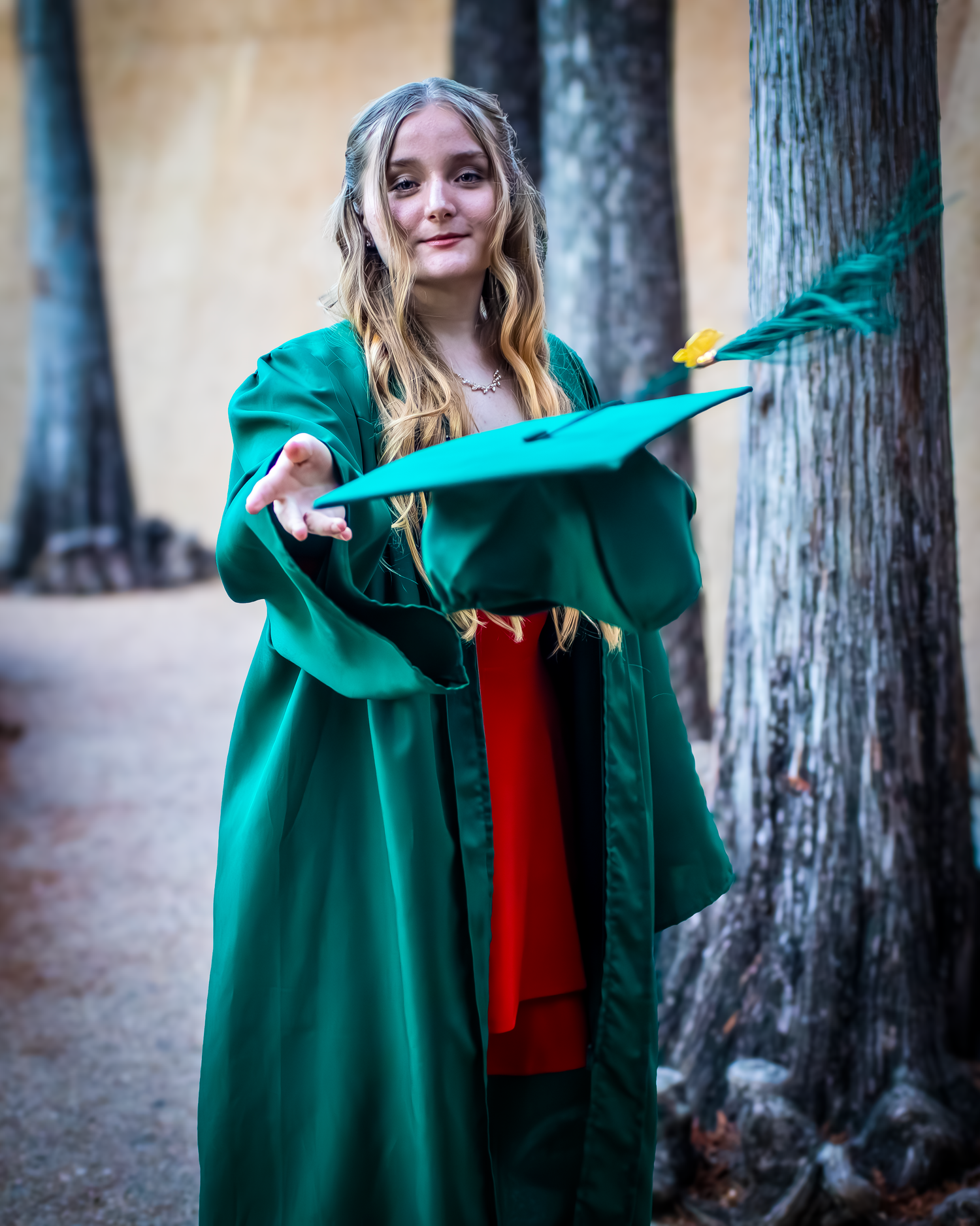 Someone wearing a bright emerald green graduation gown and red stole poses outdoors between trees during a graduation ceremony.