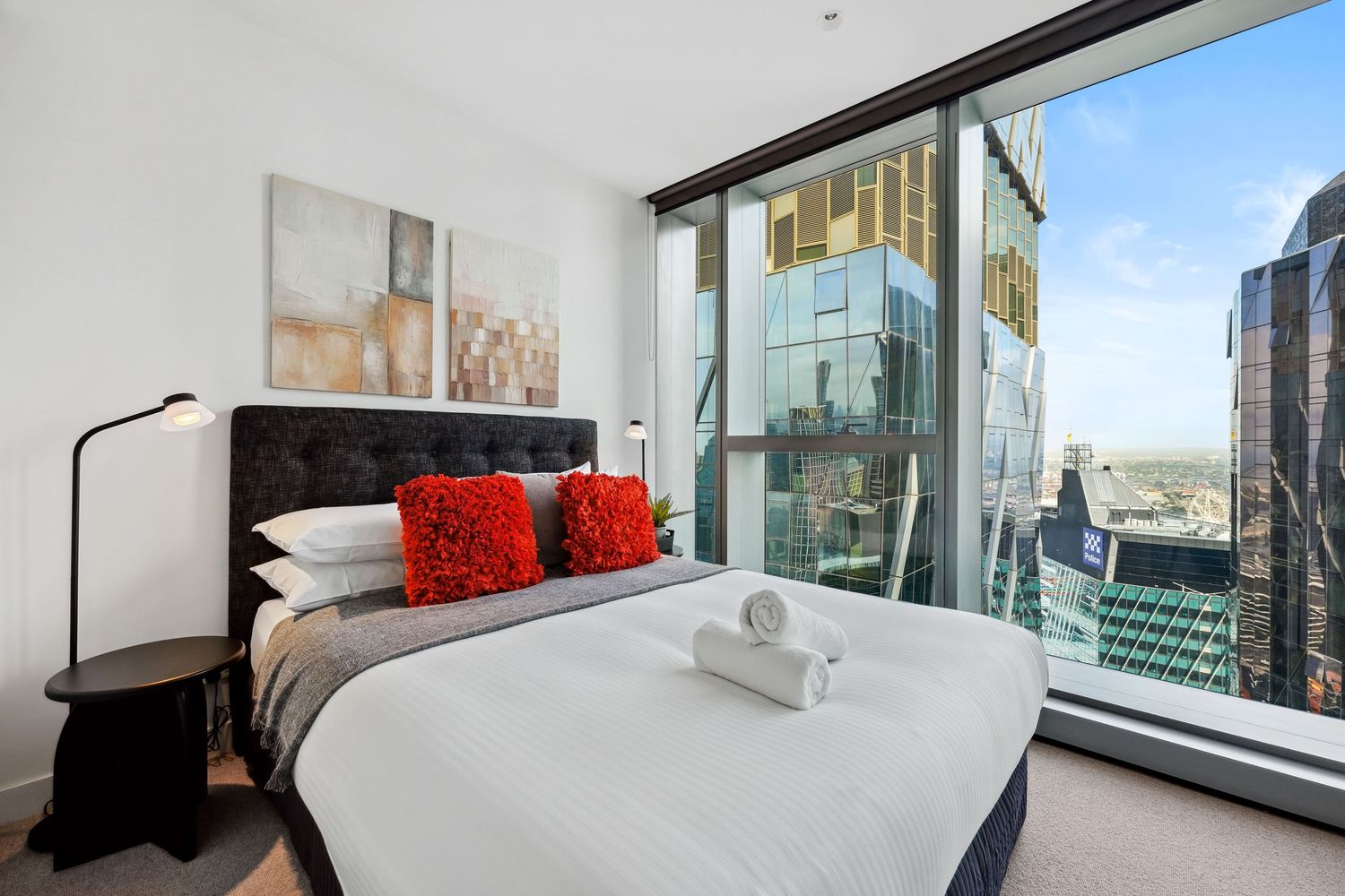 Modern apartment bedroom with floor-to-ceiling windows, city views, white bedding, red accent pillows and contemporary decor.