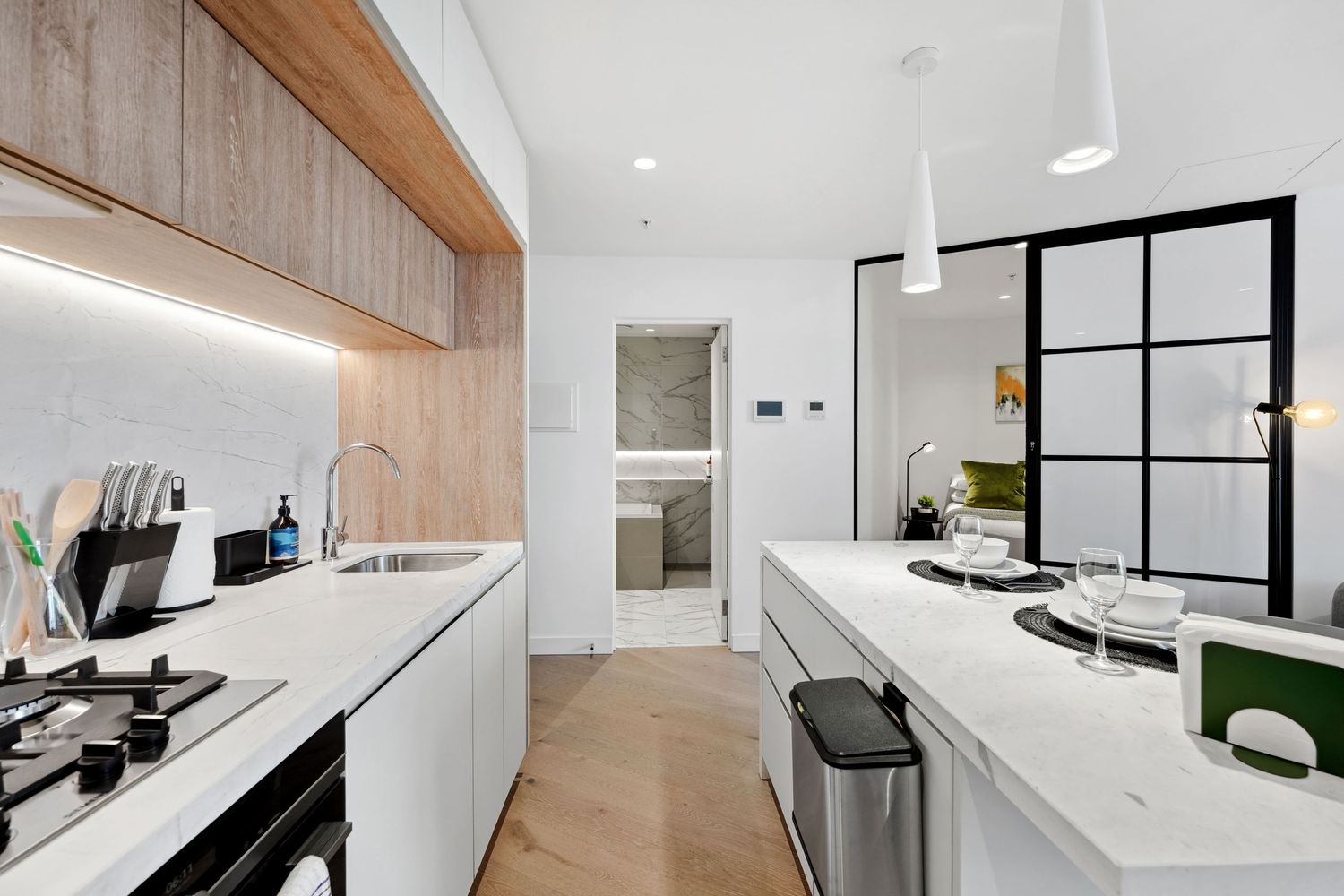 Modern white kitchen with wood accents featuring sleek countertops, gas cooktop, and industrial black-framed glass partition.