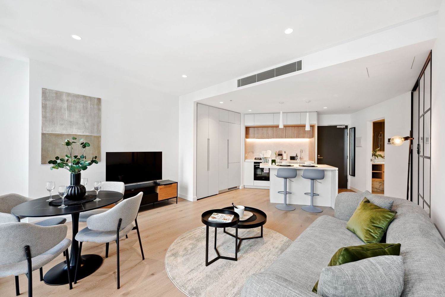 Modern open plan apartment with gray sofa, dining area, TV and white kitchen featuring contemporary minimalist design.