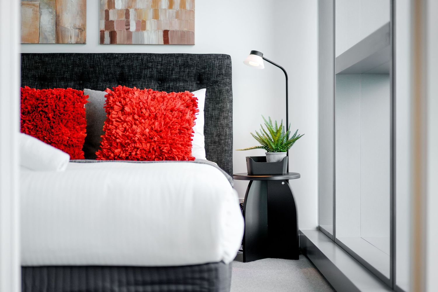 Modern bedroom interior with black headboard, crisp white bedding and vibrant red throw pillows next to a floor lamp.