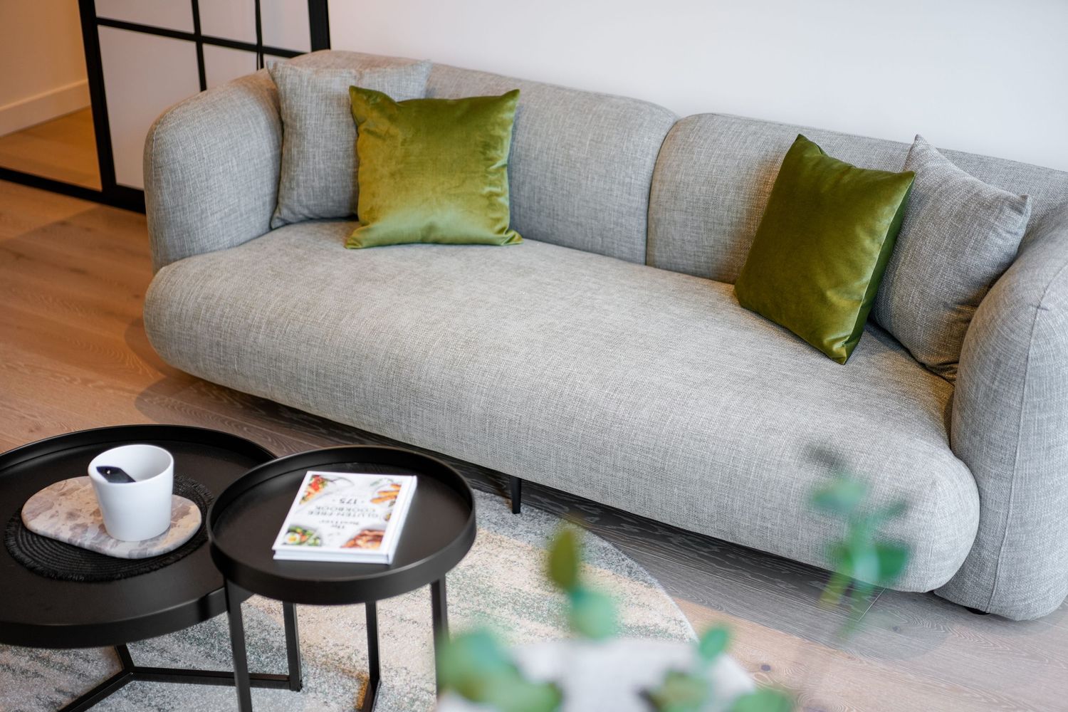 Modern gray curved sofa with green velvet accent pillows and nested black coffee tables with coffee cup and decor.