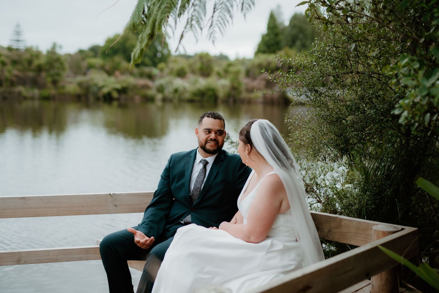 A couple sits together in a wooden boat by a serene lake during their wedding photoshoot.