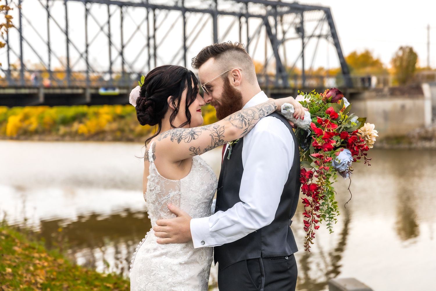 Newlywed couple embraces near waterfront with rustic bridge and autumn foliage in background.