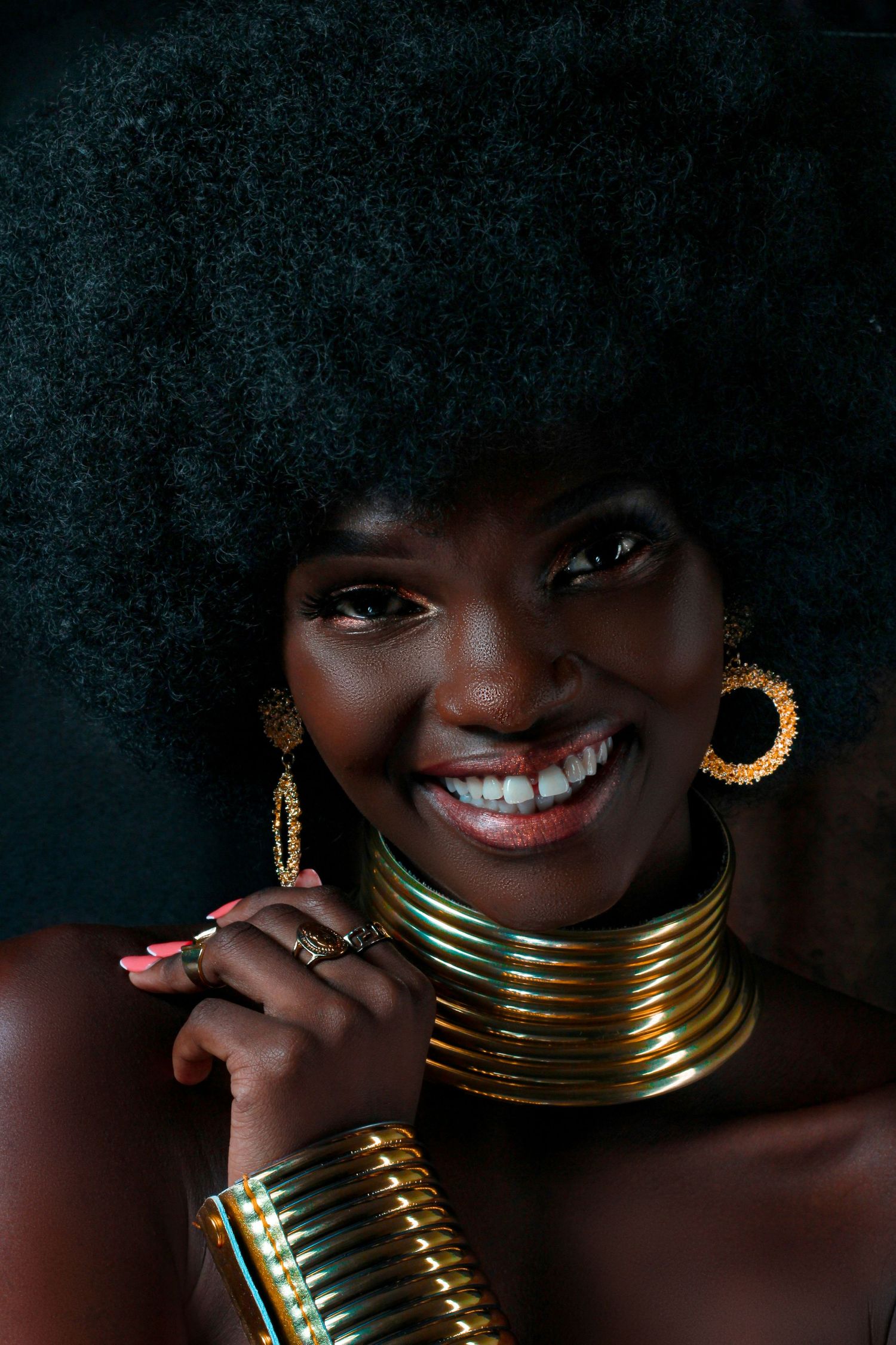 Striking beauty portrait featuring gold neck rings and hoop earrings against dark background.