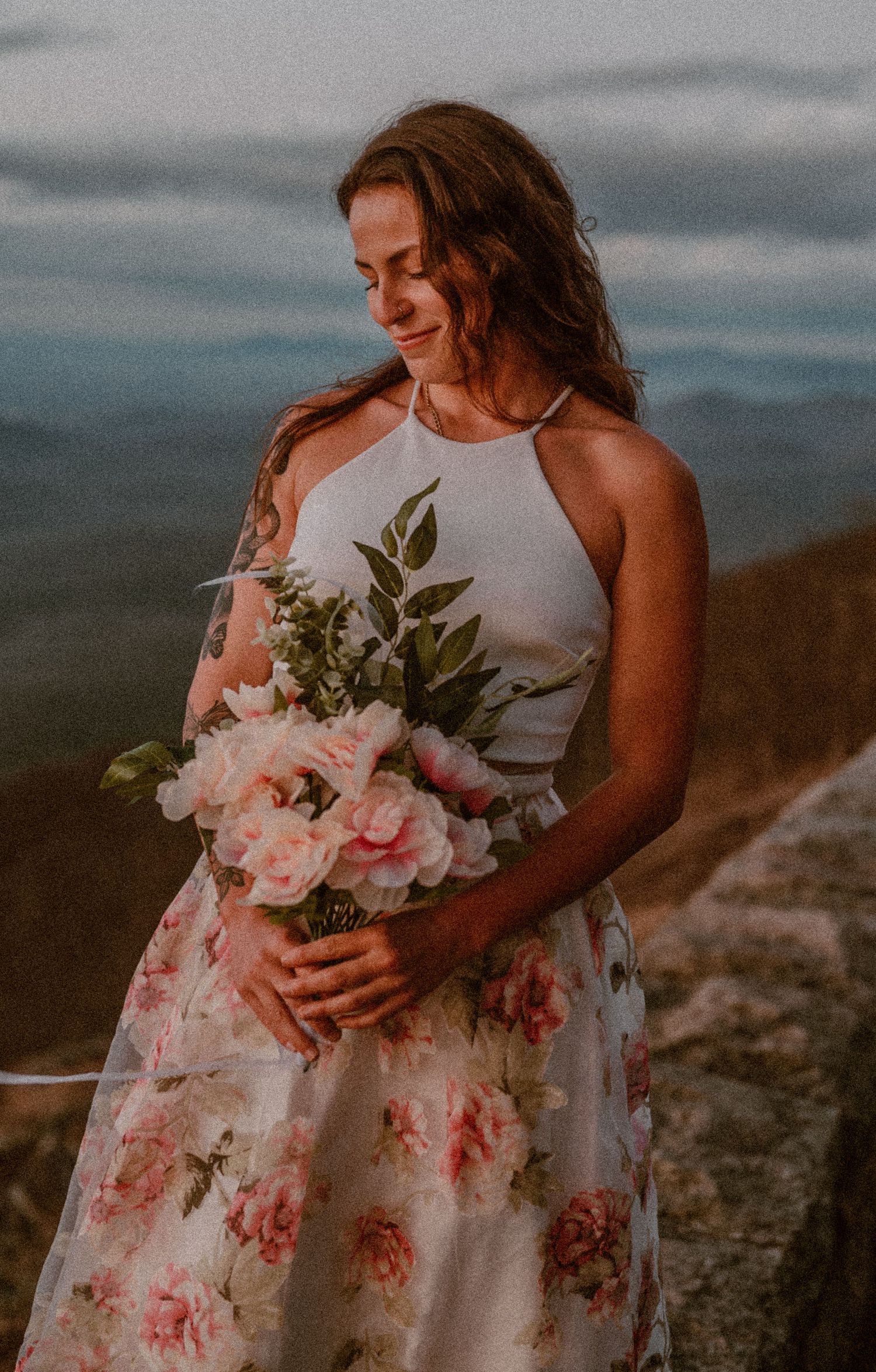 A person in a floral dress holds a bouquet of pink flowers while standing outdoors at sunset against a moody sky. photo by Samantha Campbell