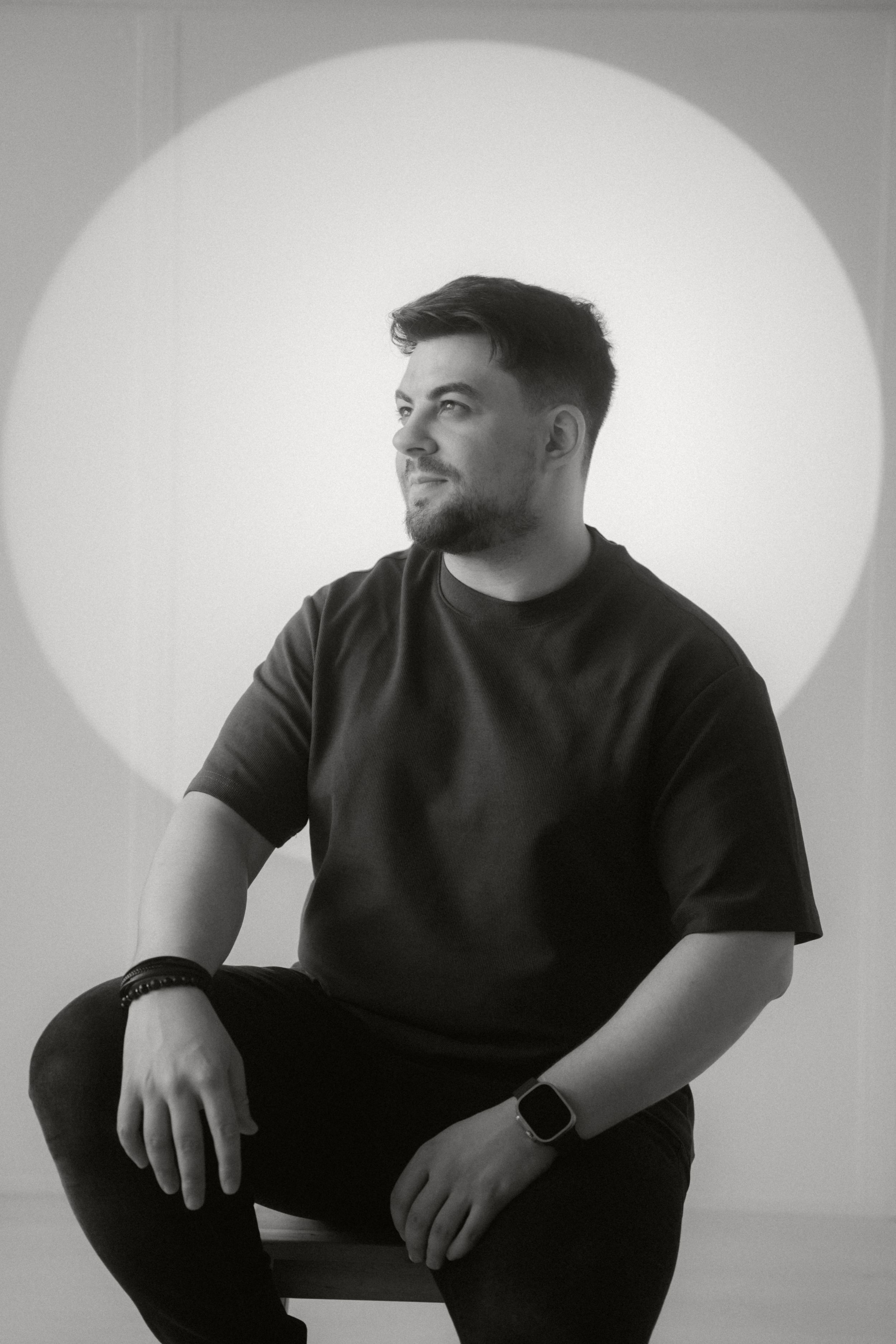 Man in dark casual attire sits on a stool against white backdrop in black and white portrait photograph.