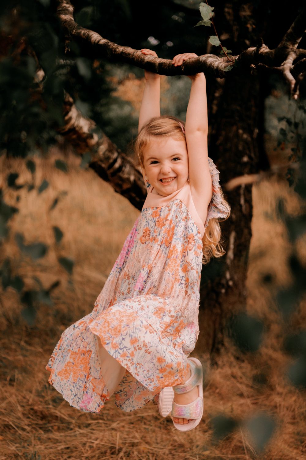 A young child in a floral dress joyfully hangs from a tree branch outdoors in warm lighting.