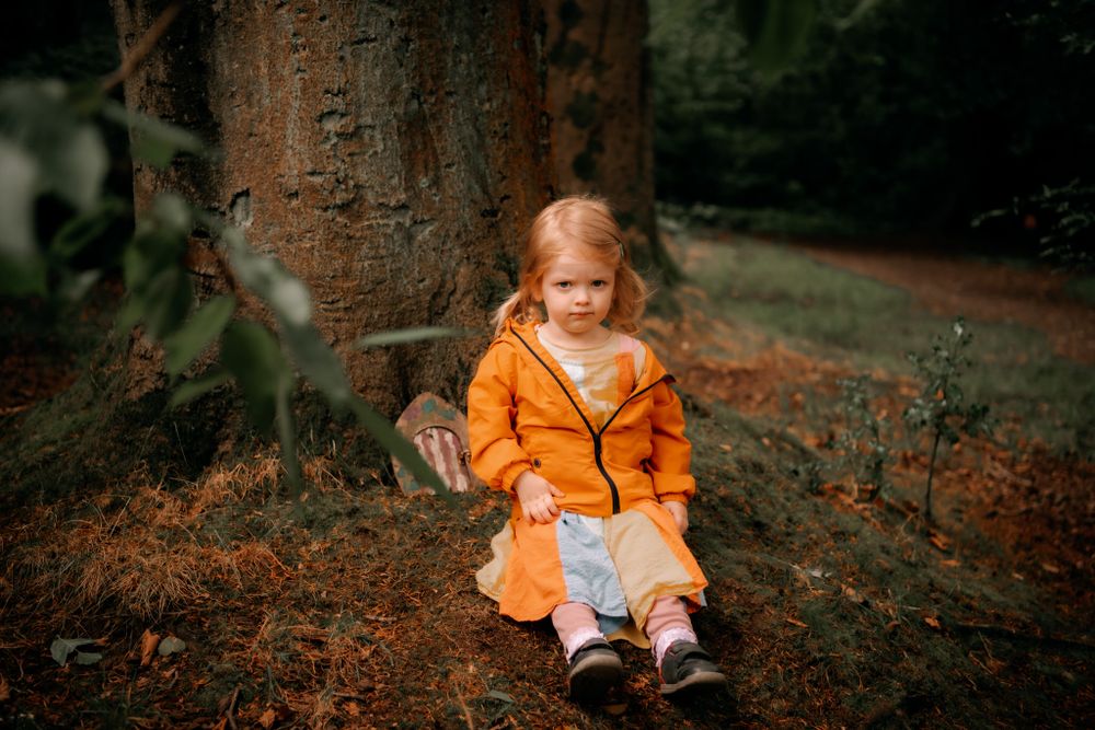 A little one wearing a bright orange coat sits on autumn leaves against a tree trunk in a forest setting.